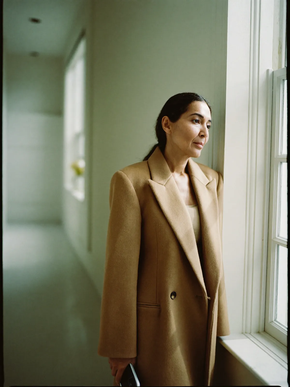 A woman in a camel coat by a window, her phone in her hand